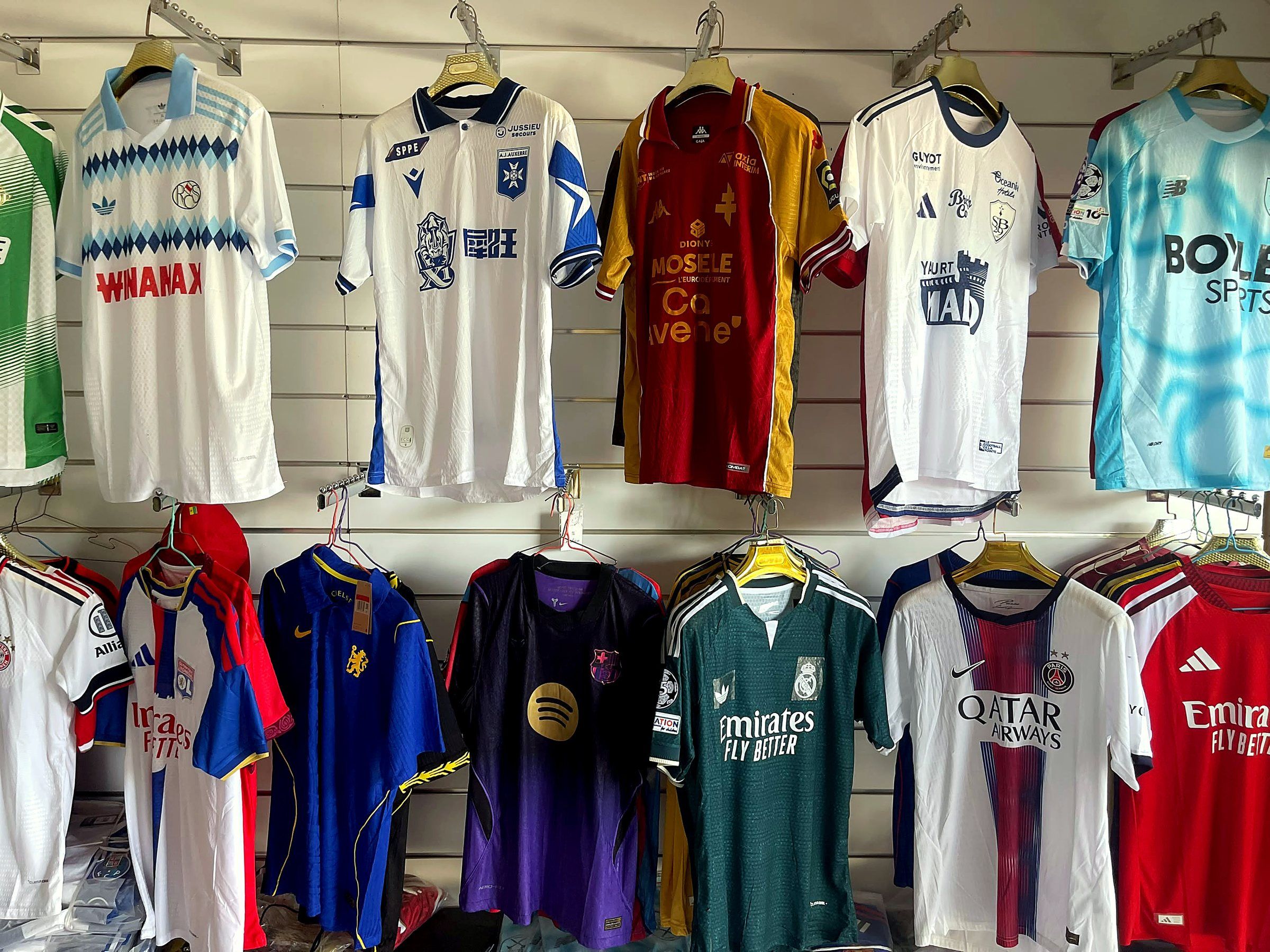 Deux rangées de maillots de clubs sur le mur blanc de la boutique, dont Auxerre, Metz, PSG et Real Madrid